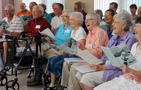 A large group of older adults seated in rows in a bright room, holding open songbooks and singing together, many of them laughing